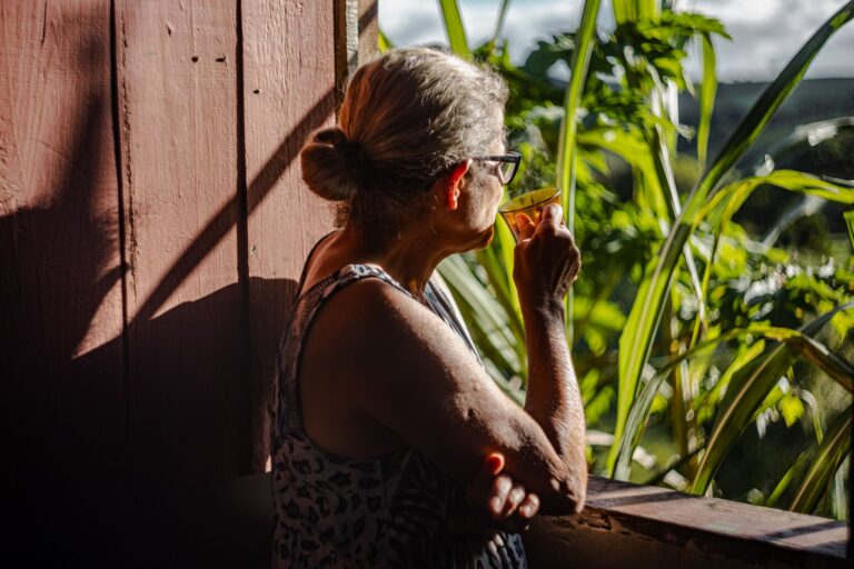 A senior woman enjoys a cup of coffee on a sunny balcony overlooking lush greenery.