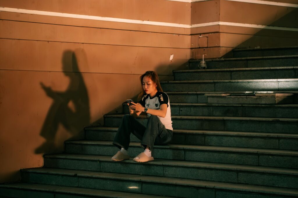 A young woman sits on steps, engaged with her phone. Warm lighting casts a distinct shadow, creating a moody urban scene.