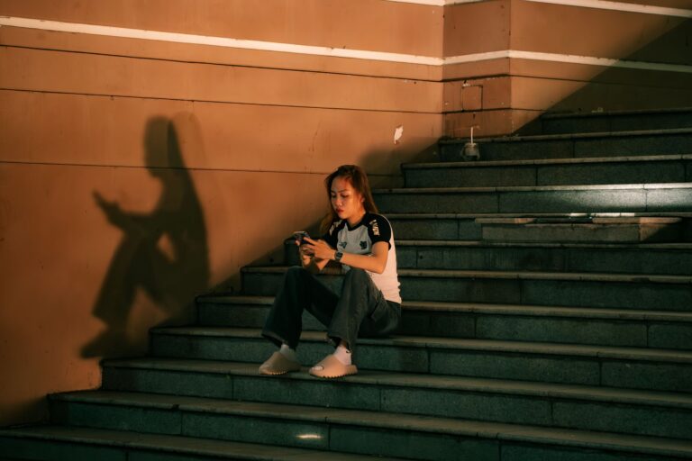 A young woman sits on steps, engaged with her phone. Warm lighting casts a distinct shadow, creating a moody urban scene.