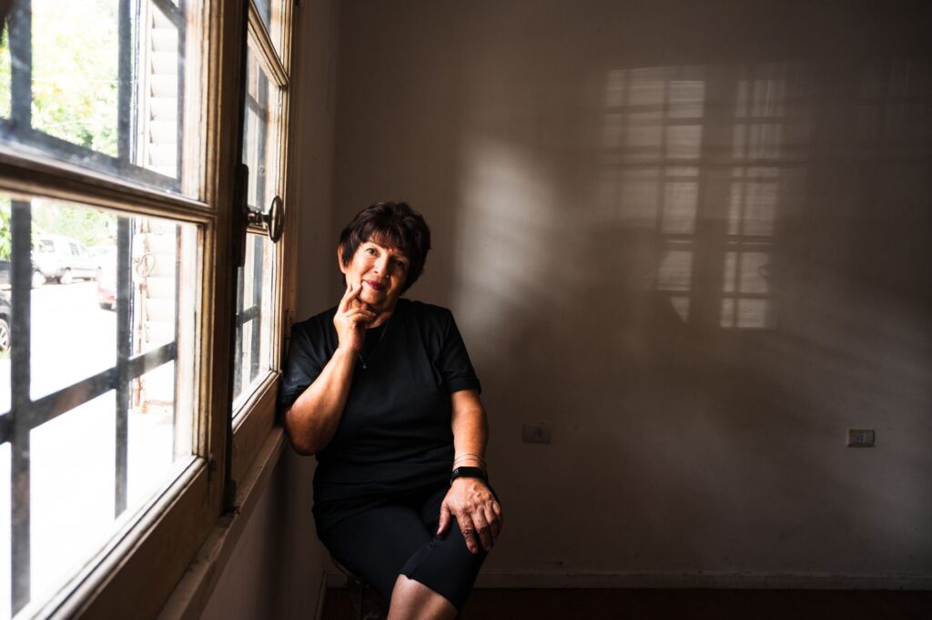 A senior woman seated indoors by a window, deep in thought, with sunlight casting shadows.