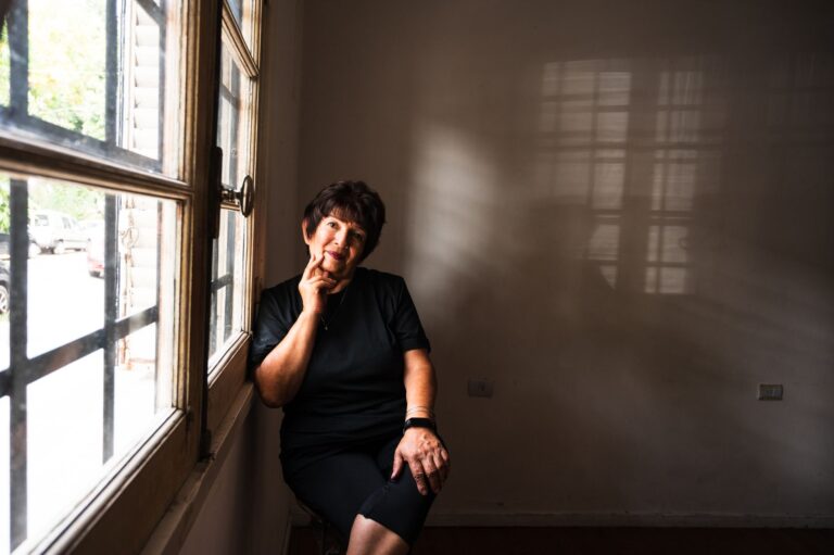 A senior woman seated indoors by a window, deep in thought, with sunlight casting shadows.