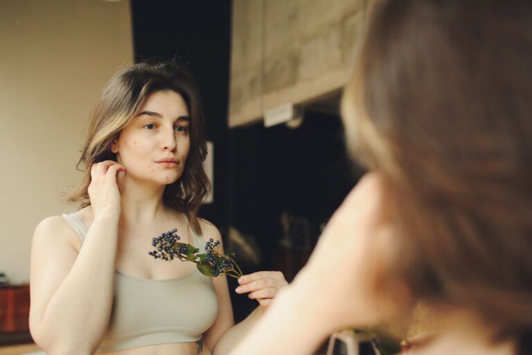 Young woman in crop top gazes at reflection, holding purple flowers indoors.