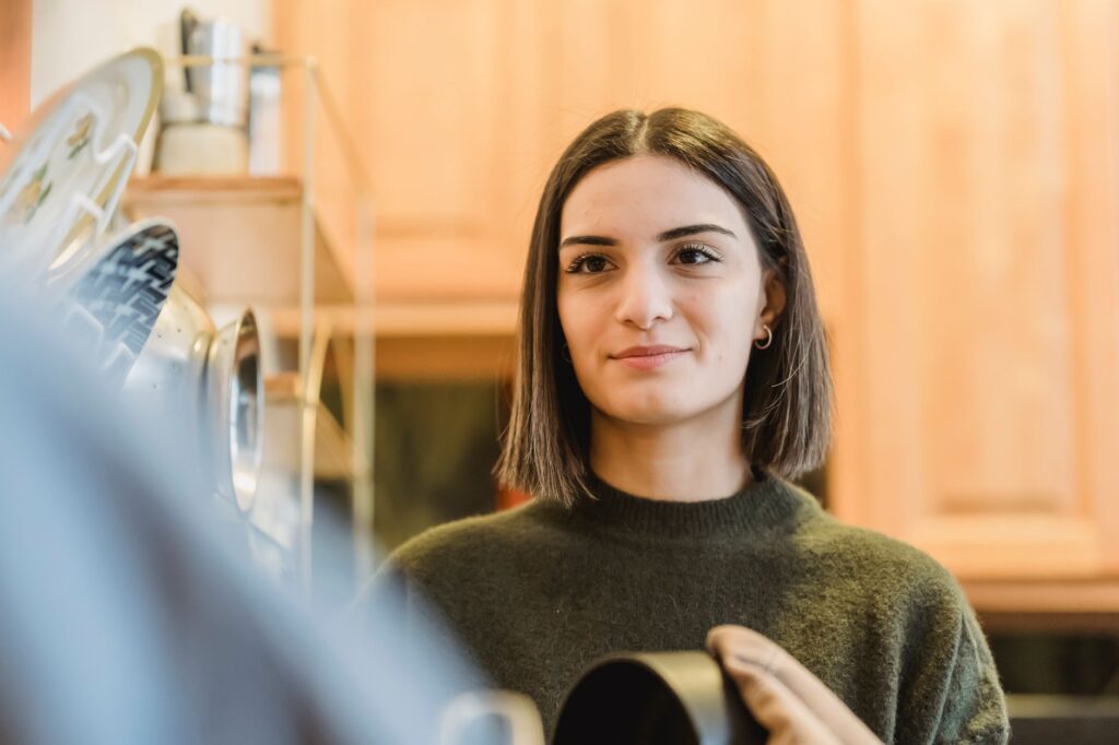Cheerful young female smiling and cleaning saucepan with textile in kitchen with plates and kitchenware