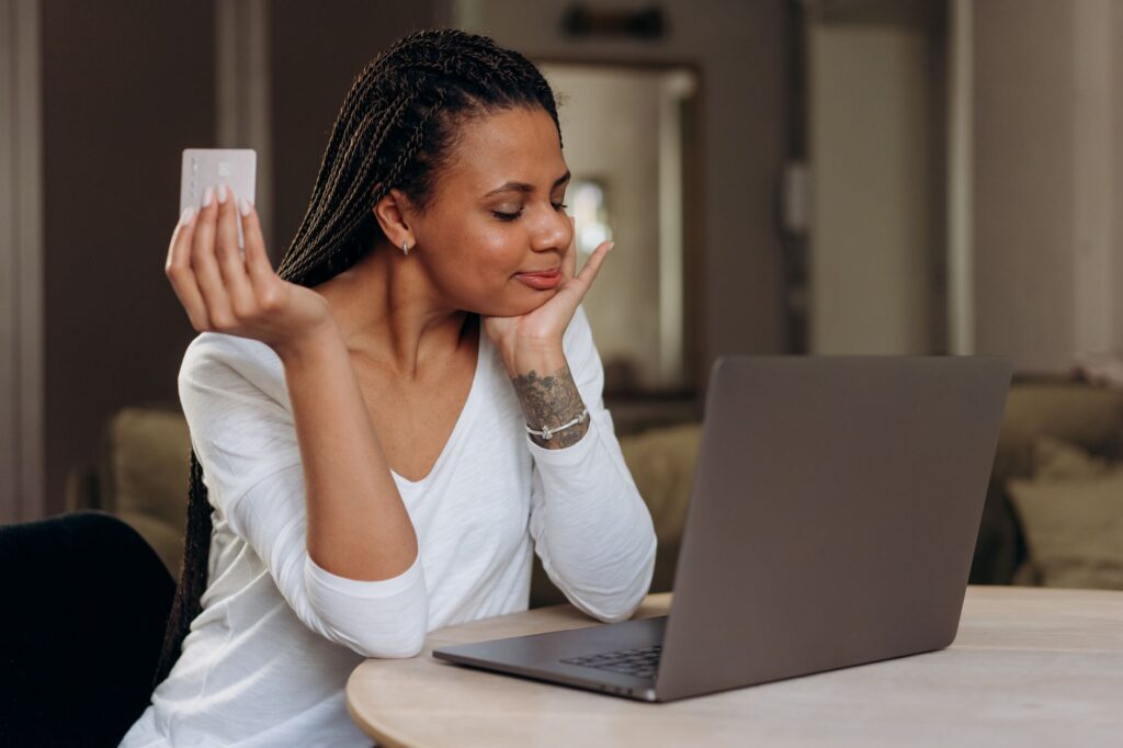 A woman enjoys shopping online, holding a credit card next to her laptop at home.