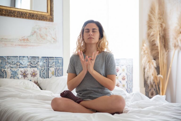 A woman meditating in a peaceful bedroom setting with soft natural light.