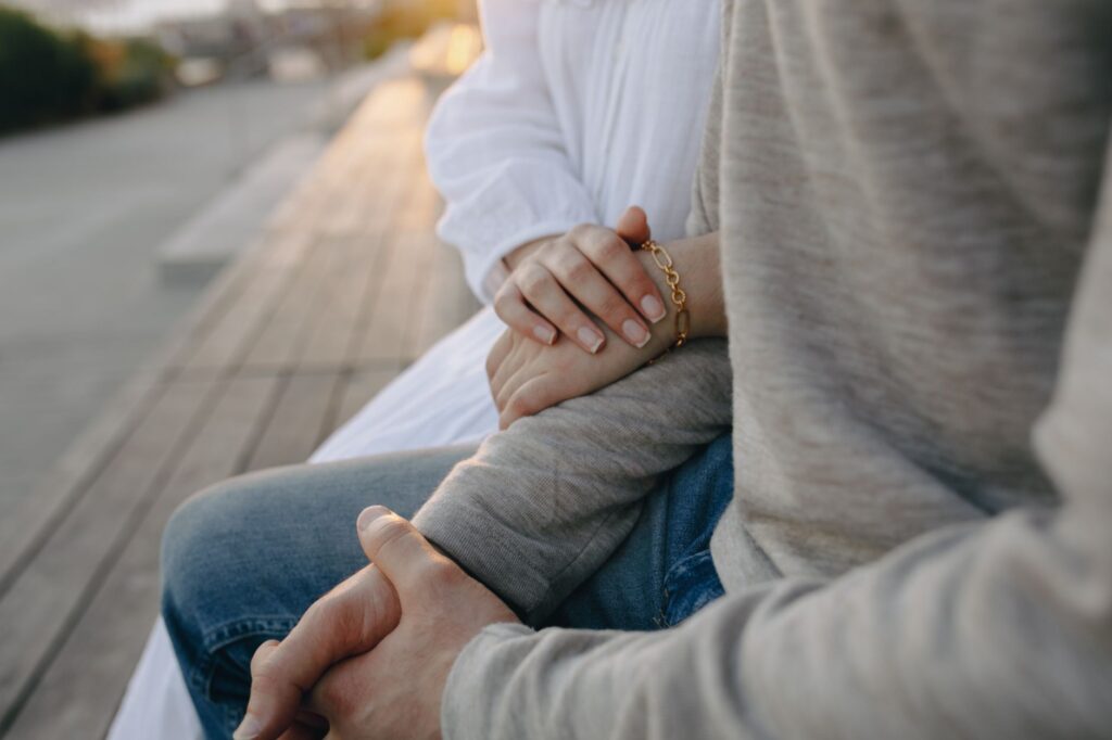 Close-up of a couple holding hands, seated on an outdoor bench during the day.