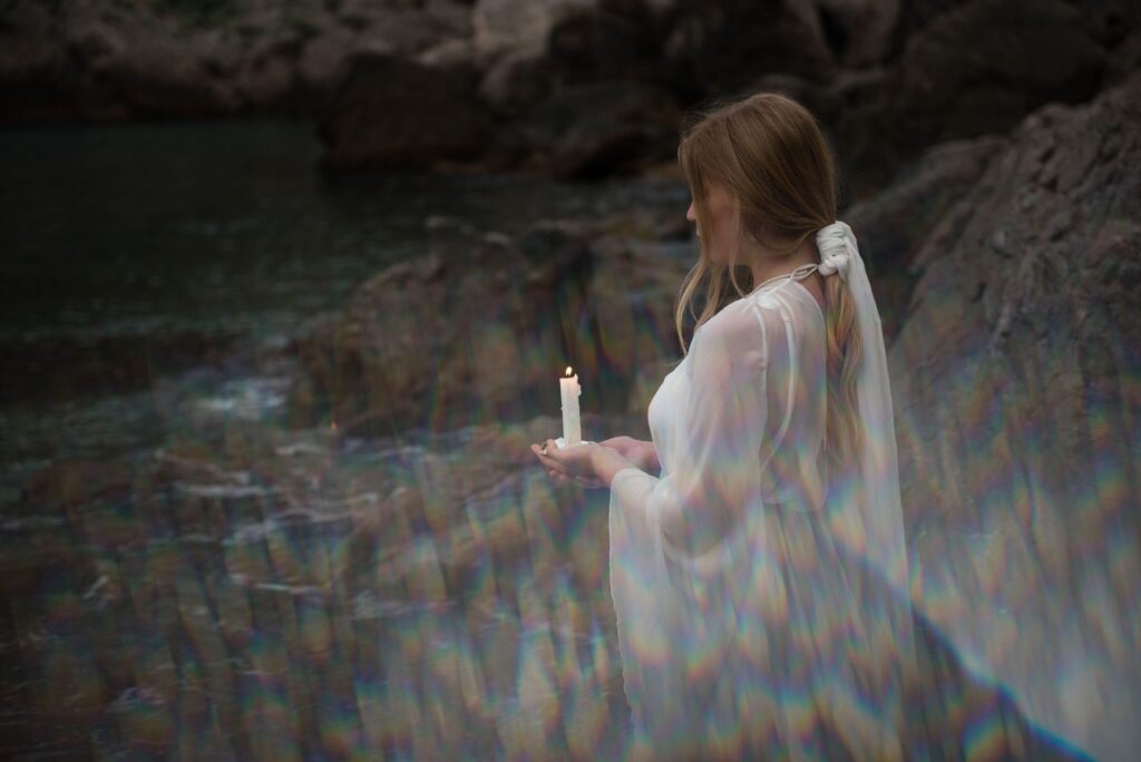 A woman in a white dress holding a lit candle beside a tranquil rocky stream.