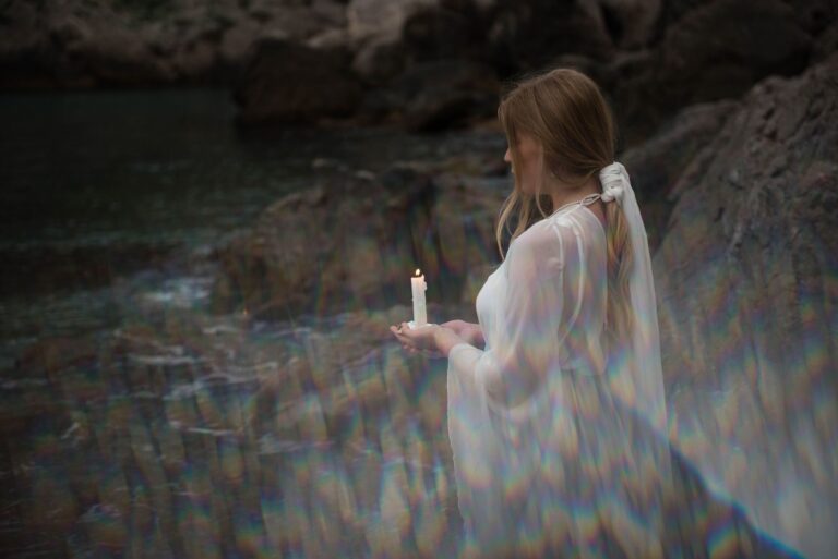 A woman in a white dress holding a lit candle beside a tranquil rocky stream.